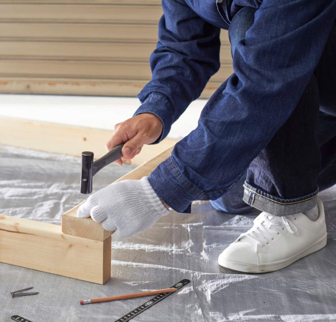 carp-2 Person wearing gloves and jeans hammering a nail into wooden planks on a covered floor.
