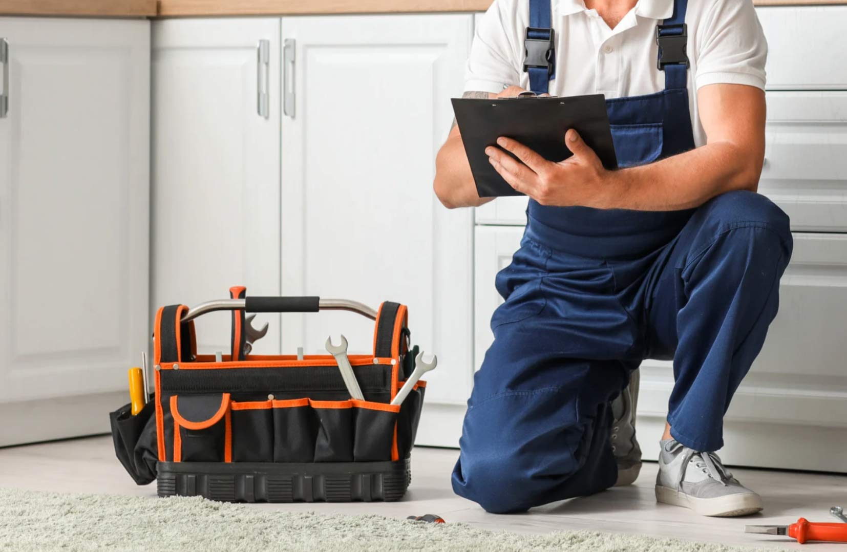 general-handy-2 A repair worker in overalls kneels with a clipboard beside a tool bag in a kitchen.