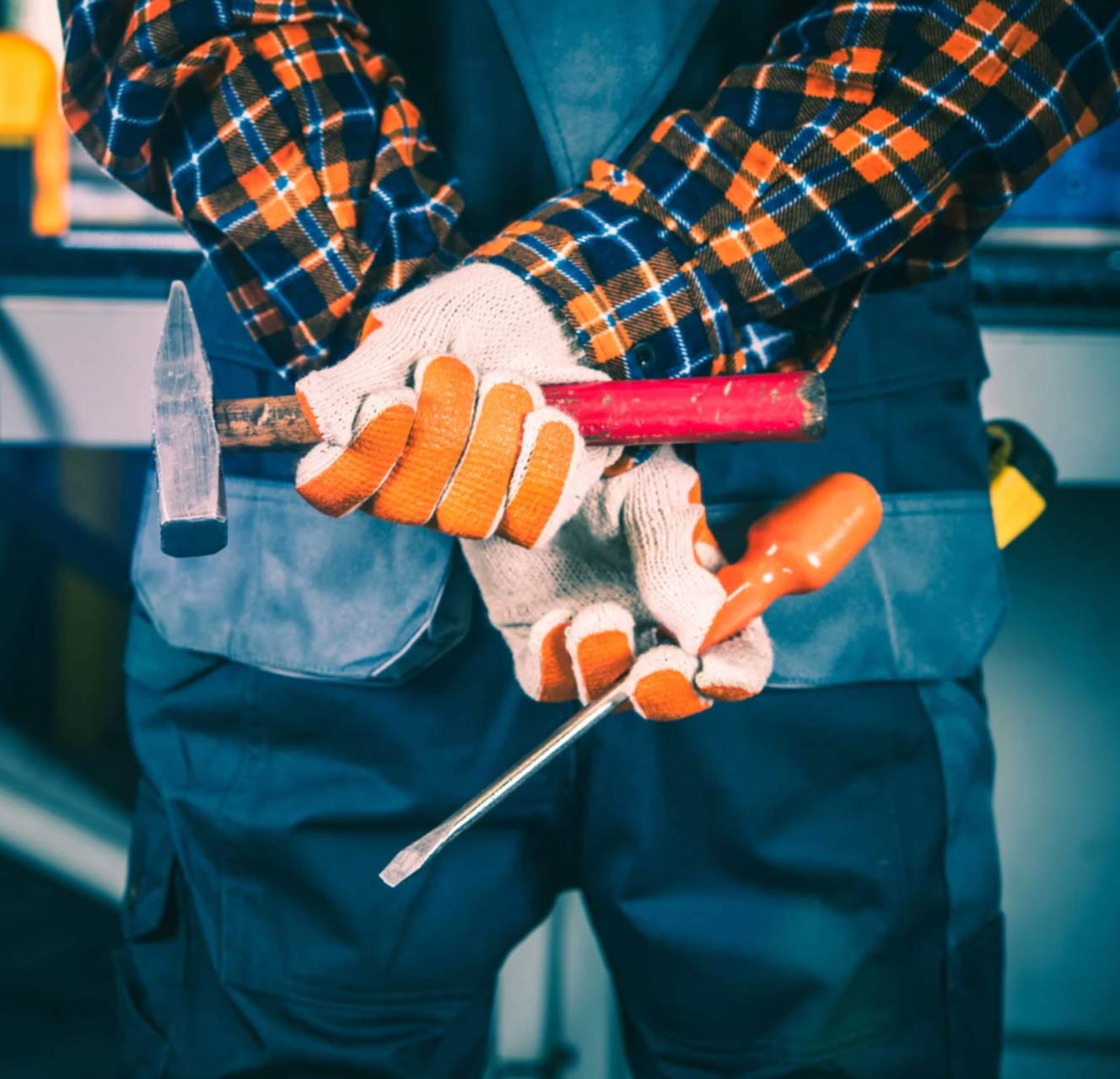 handy-img Person in work gloves holds a hammer, screwdriver, and chisel, wearing a plaid shirt and overalls.