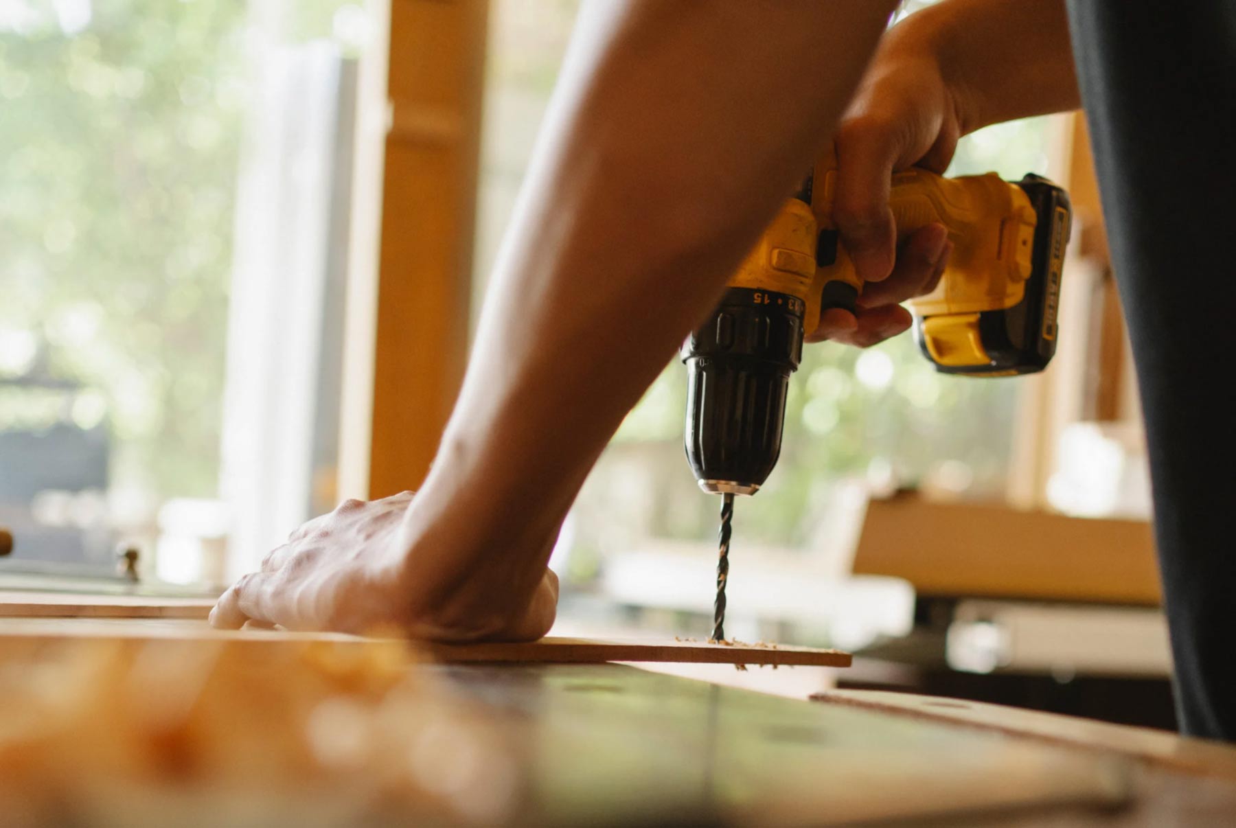 handy-img Person using a yellow power drill to make a hole in a wooden board on a workbench.