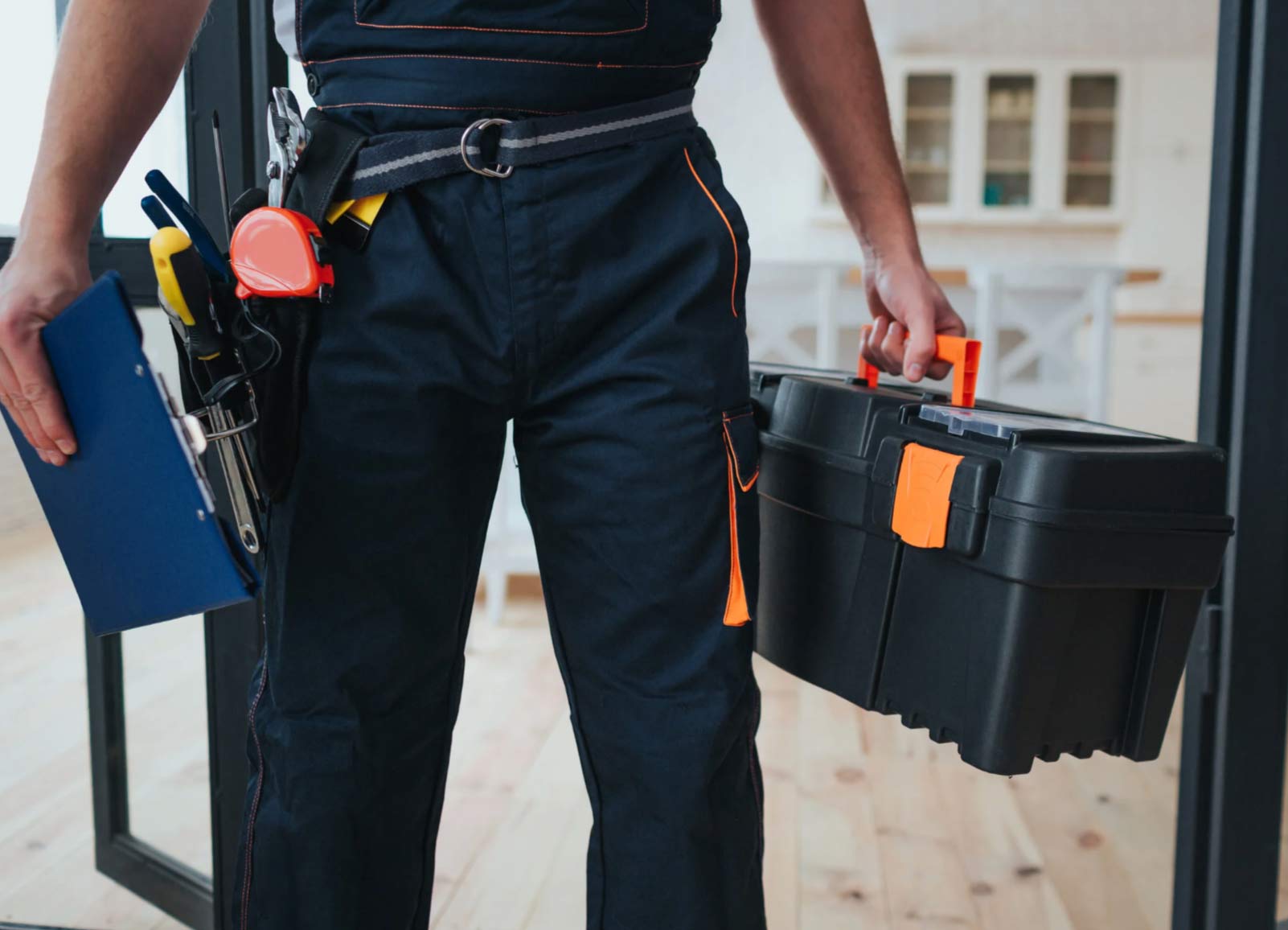 handyman-img A worker in overalls holds a toolbox and a folder, with tools attached to a belt around his waist.