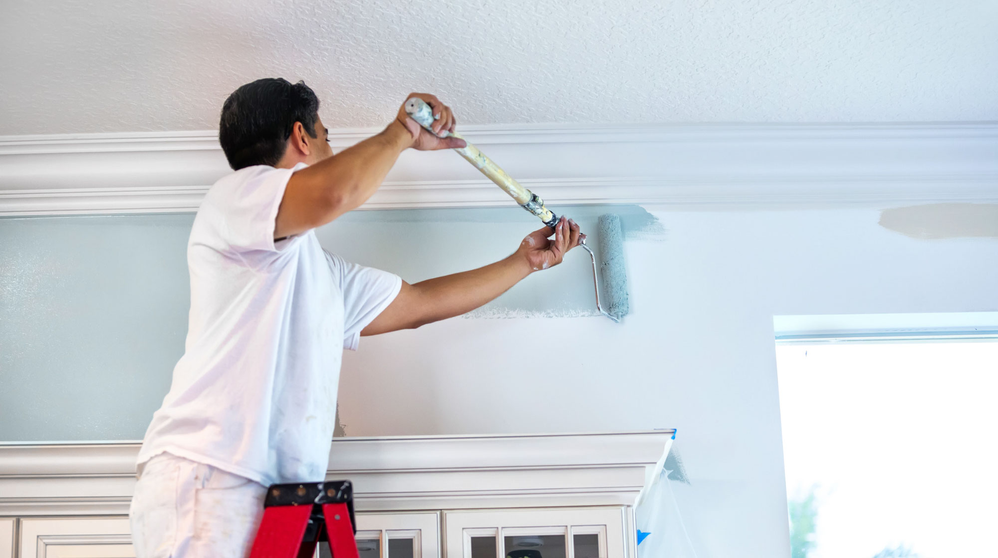 painting-us Person on a ladder painting a wall light blue near the ceiling in a bright room.