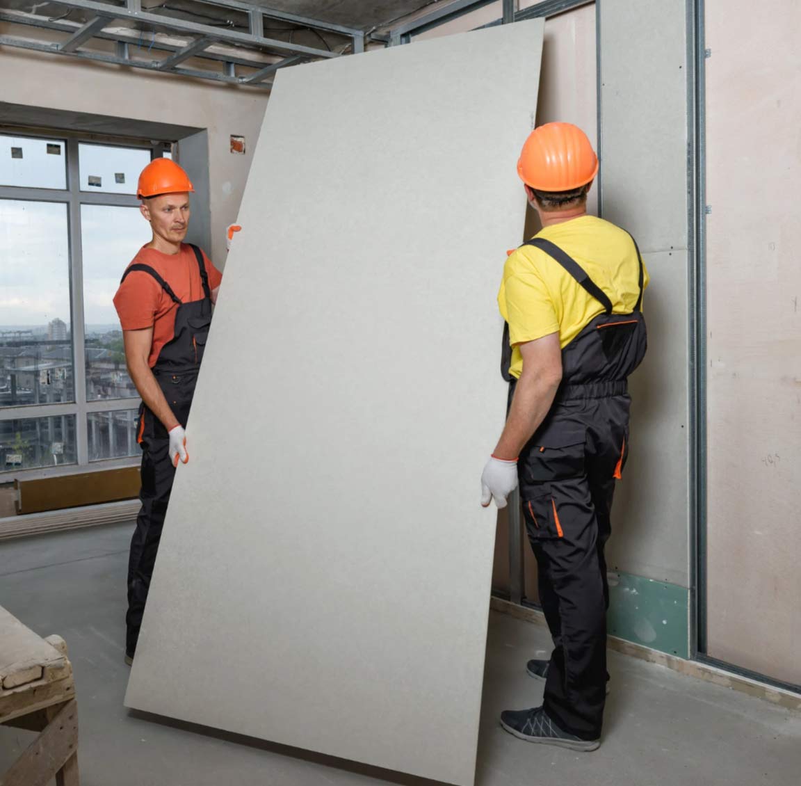 sheet-2 Two construction workers in hard hats carry a drywall sheet inside a building under construction.