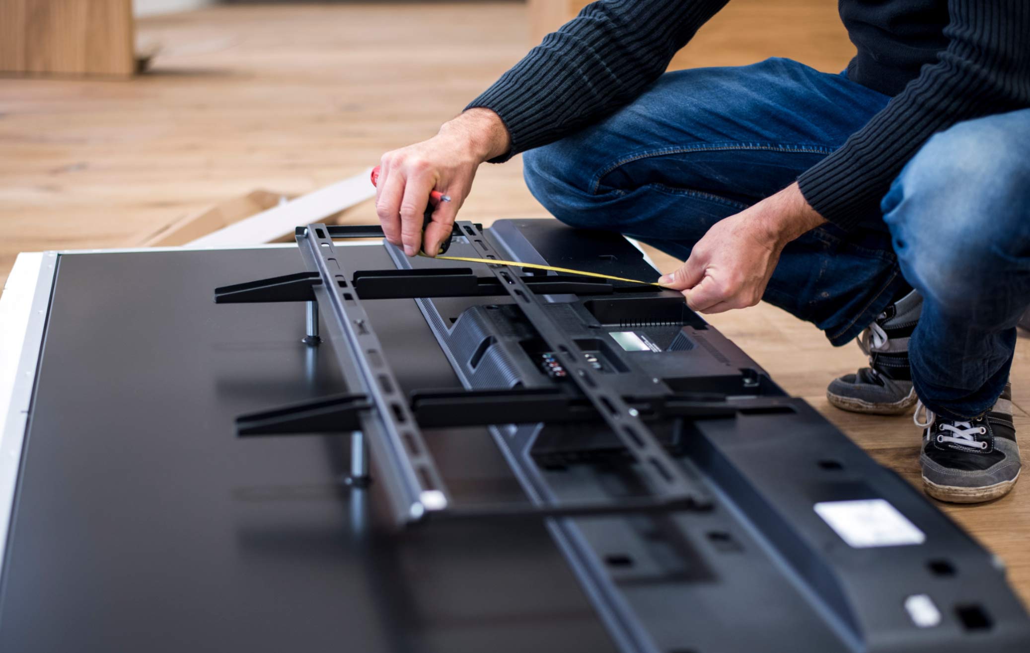 tv-1 Person kneeling and measuring a TV mount bracket on the back of a flat-screen television.